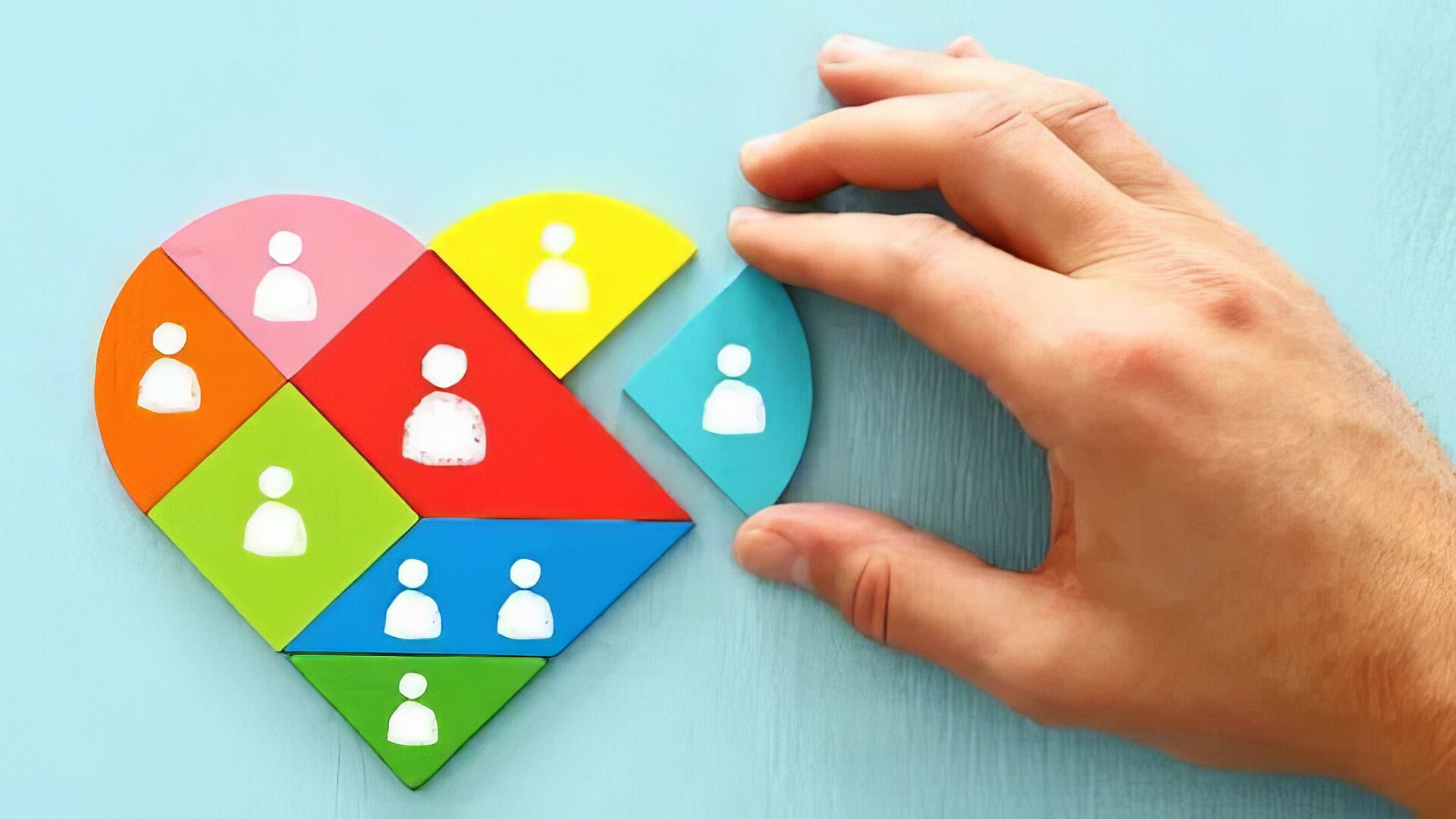 Holzpuzzles in Herzform welches durch eine dargestellte Hand zusammengesetzt wird. Jedes Puzzlesteil ist in unterschiedlicher Farbe und mit einer Illustration eines Menschen dargestellt.