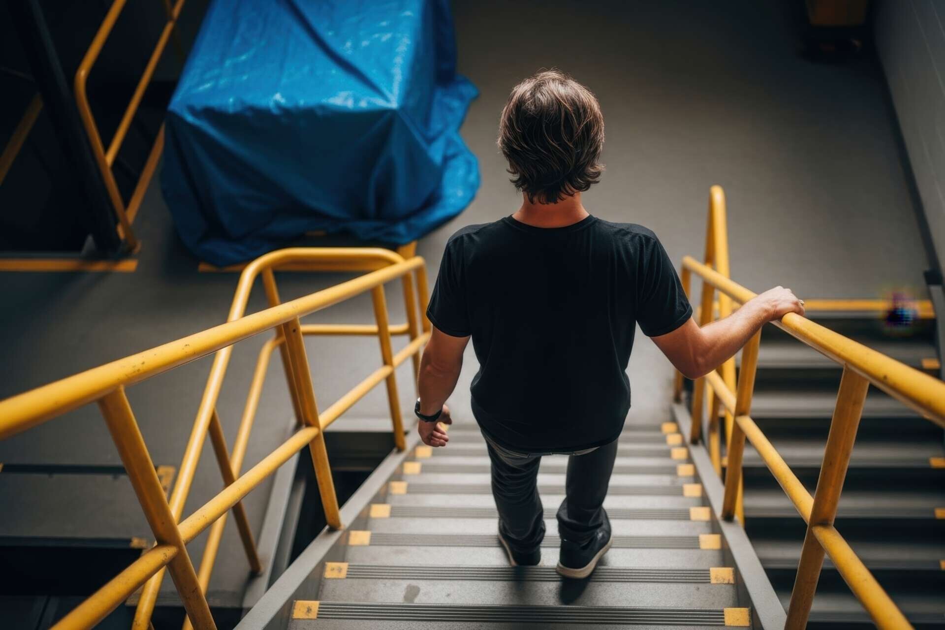 Rückansicht einer Person, die eine Industrietreppe mit leuchtend gelben Geländern in einer Lagerhalle oder Fabrik hinuntergeht 