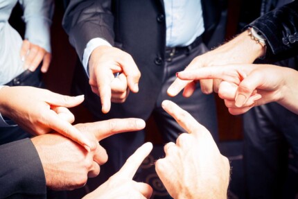 Händen verschiedener im Kreis stehender Menschen, deuten mit dem Zeigefinger aufeinander 