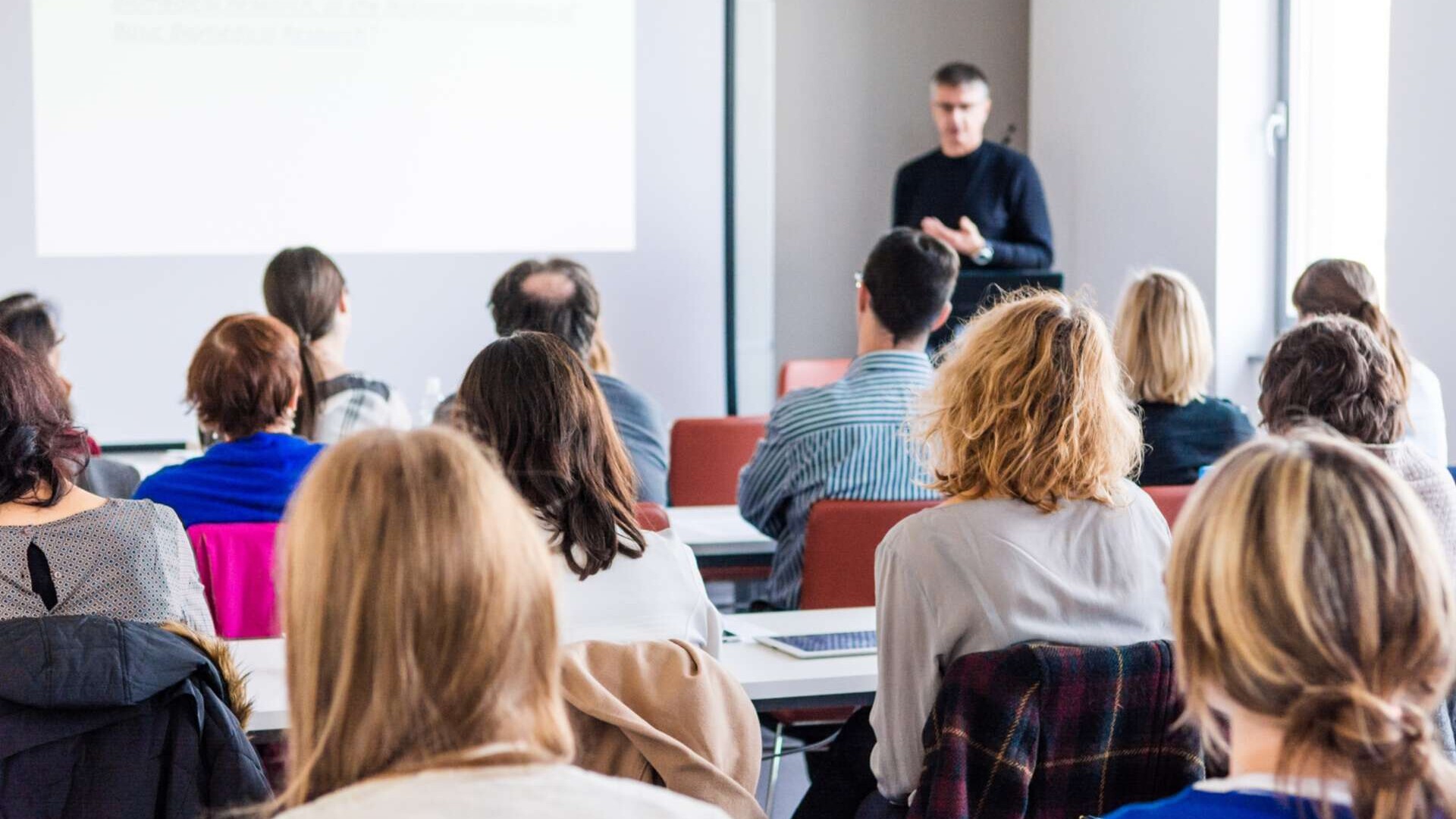 Seminarsituation in Präsenz, Dozent im Hintergrund und Teilnehmende von Hinten