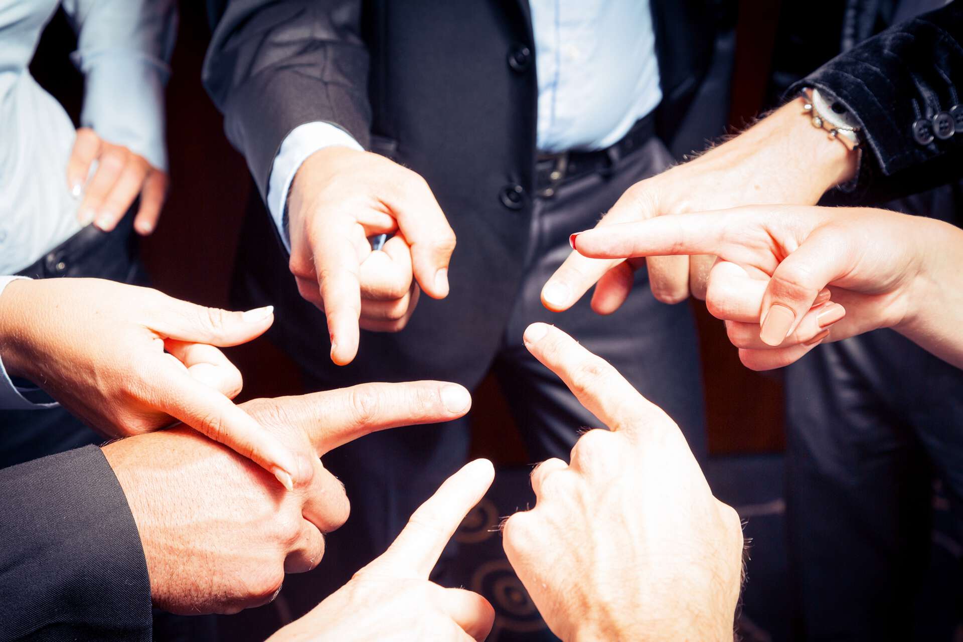 Händen verschiedener im Kreis stehender Menschen, deuten mit dem Zeigefinger aufeinander 