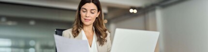 Frau sitzt am Laptop mit Papier in der Hand
