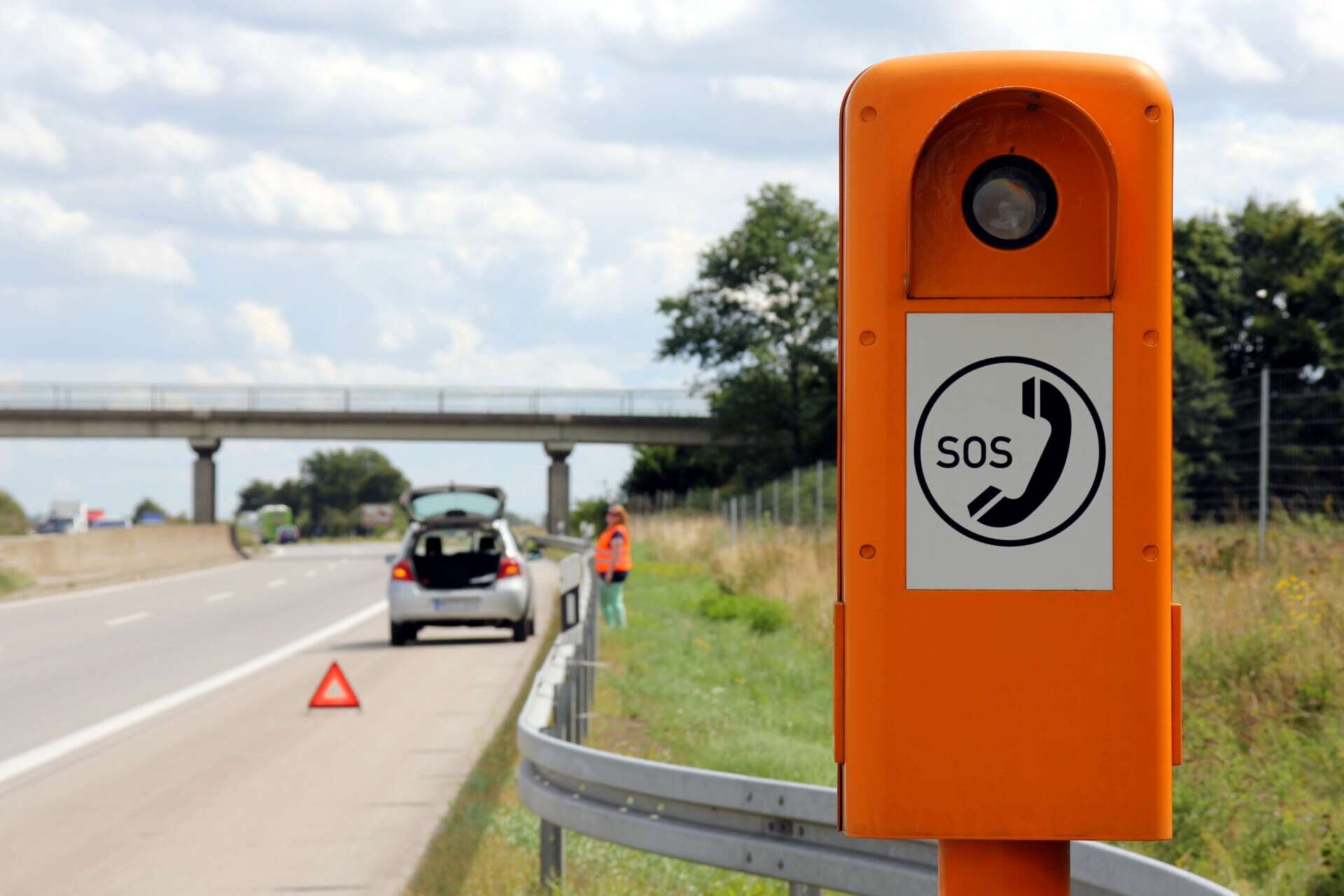 Frau mit Warnweste hinter der Leitplanke auf der Autobahn. Im Vordergrund ist ein Warndreieck und eine Notrufsäule.