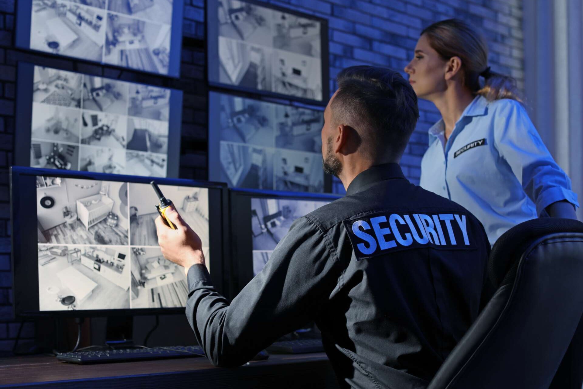 Zwei Beschäftigte im Sicherungsdienst schauen  auf einem Monitor mit Aufnahmen von Überwachungskameras.