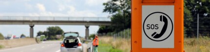 Frau mit Warnweste hinter der Leitplanke auf der Autobahn. Im Vordergrund ist ein Warndreieck und eine Notrufsäule.