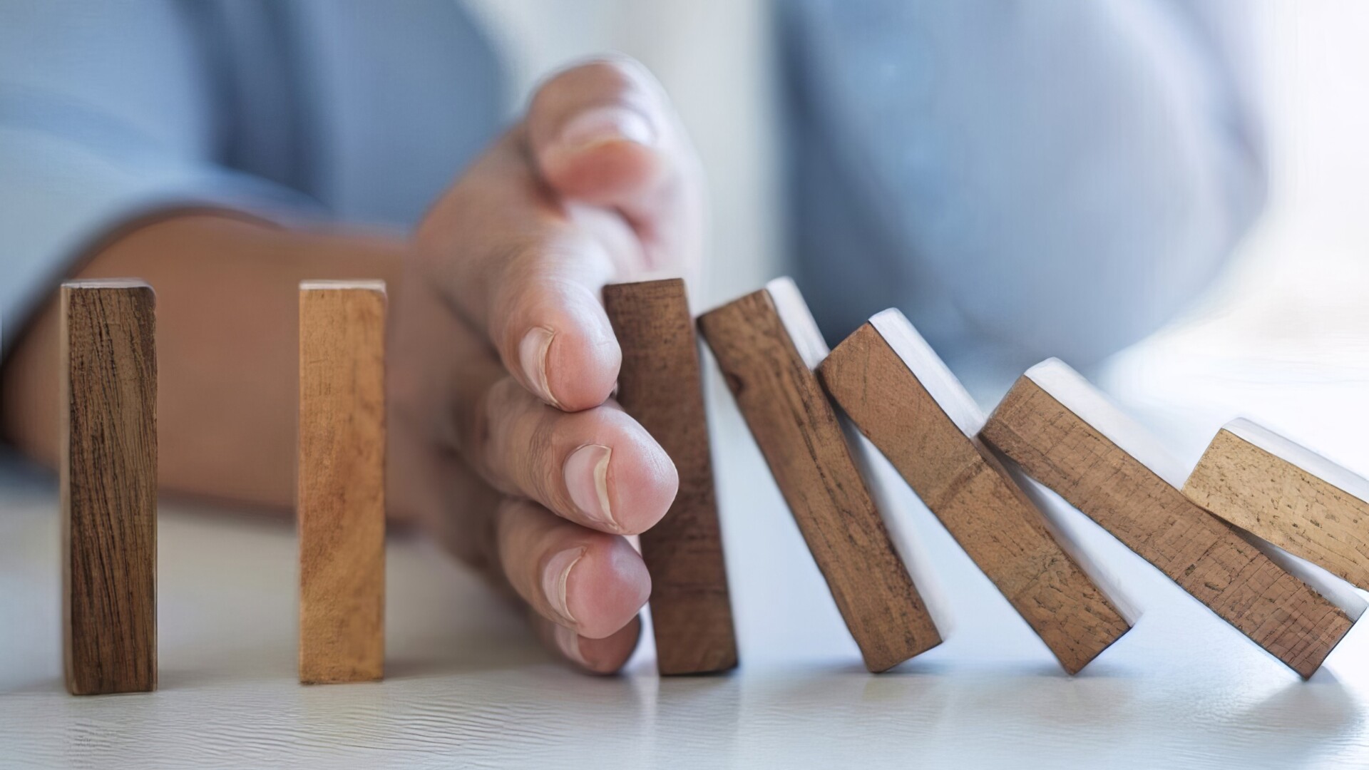 Umfallende Holzdominosteine die von einer dazwischenliegenden Hand aufgehalten werden.