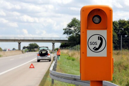 Frau mit Warnweste hinter der Leitplanke auf der Autobahn. Im Vordergrund ist ein Warndreieck und eine Notrufsäule.