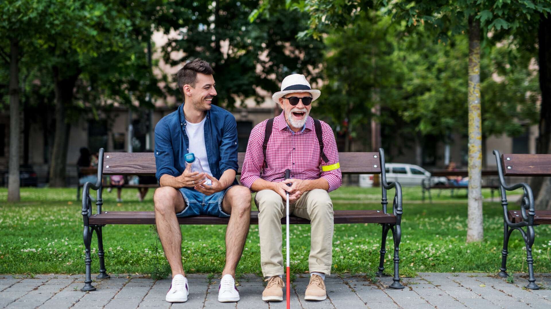 Zwei Menschen sitzen auf einer Parkbank und lachen. Einer hat einen Blinenstock und eine Sonnenbrille auf